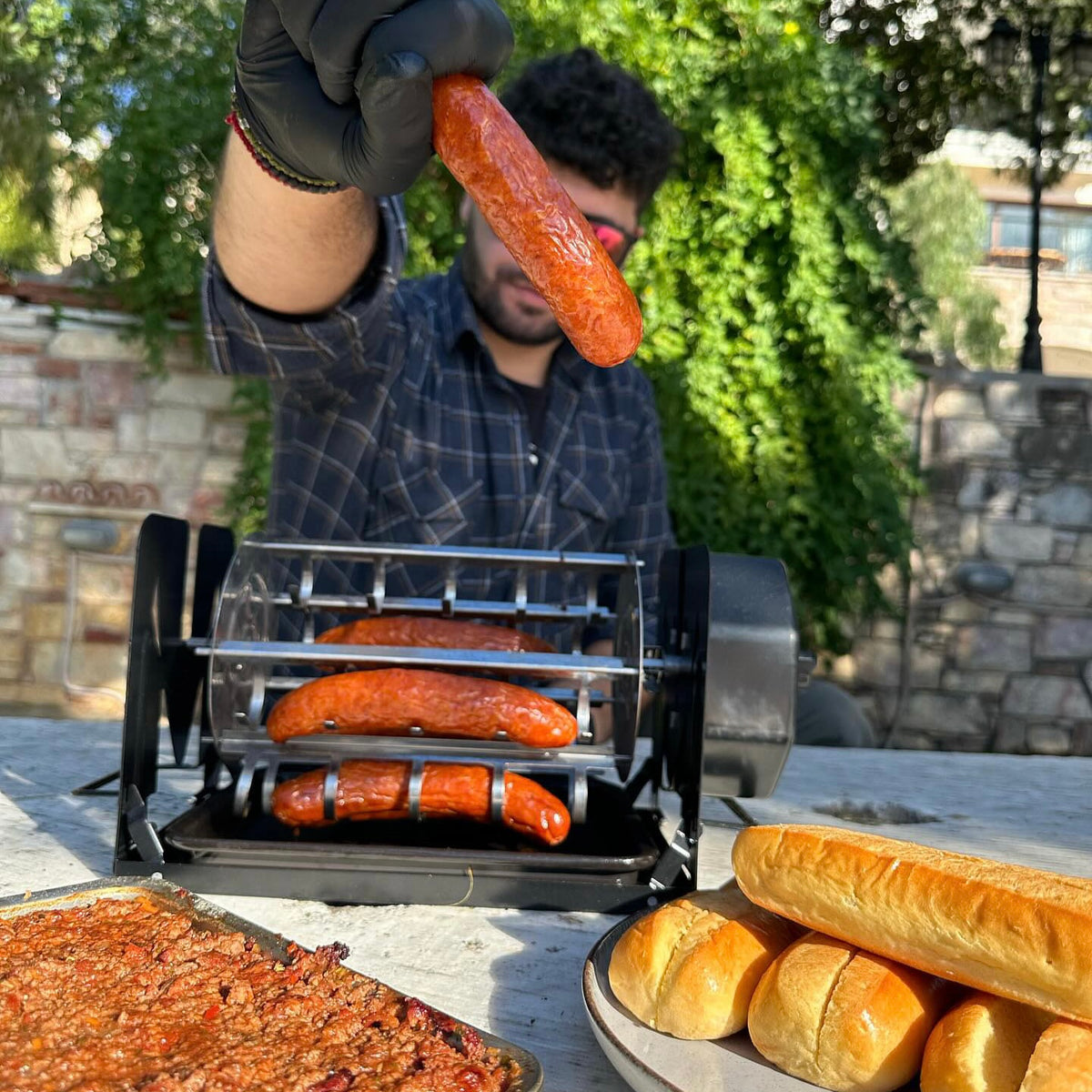 A person holding a cooked sausage above a black ROTO-Q 360 hot dog rotisserie cradle