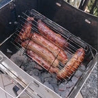 ROTO-Q 360 Rotisserie Cage (Flat) filled with sausages grilling over hot charcoal coals.