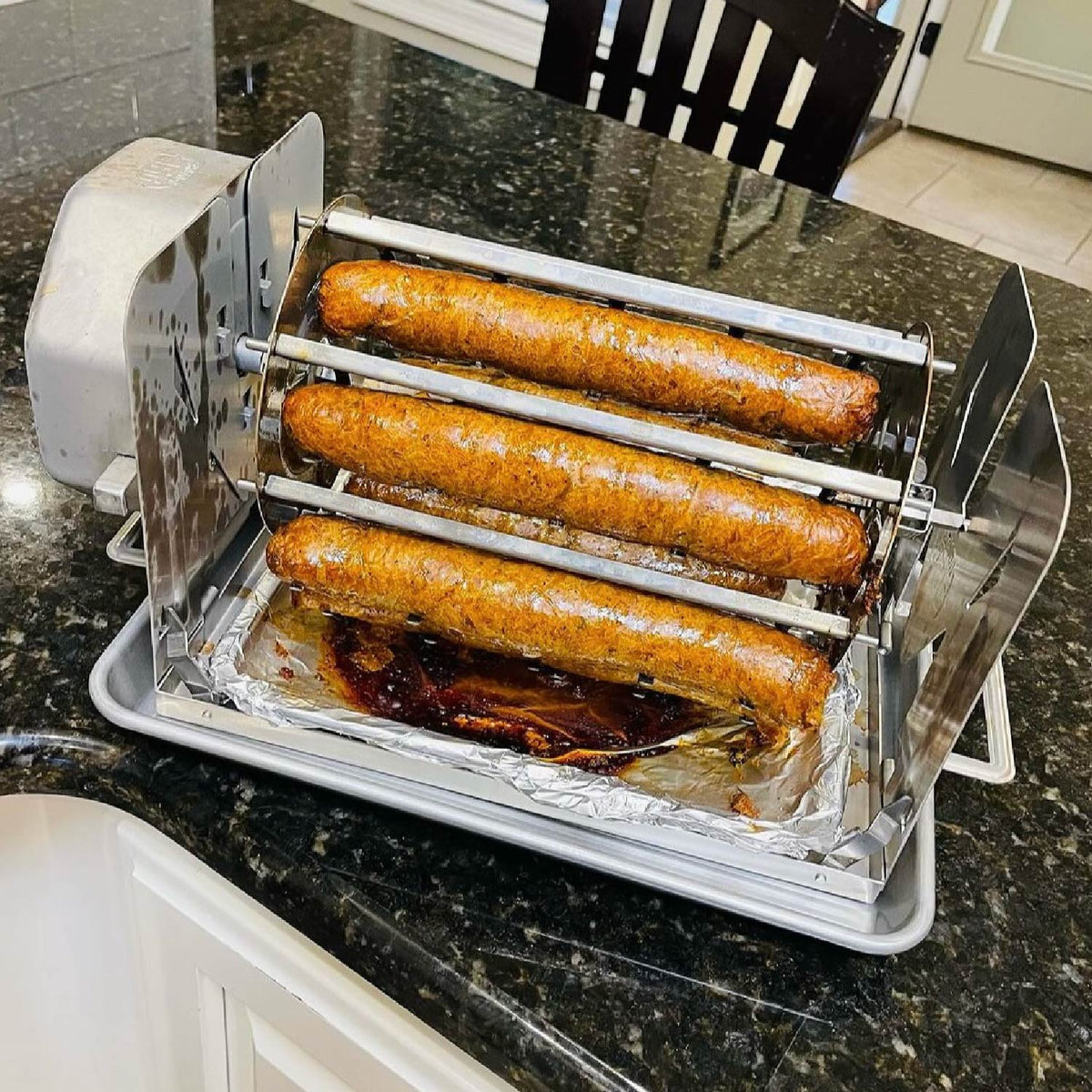 ROTO-Q 360 Sausage Rotisserie holding cooked sausages on a kitchen counter with a foil-lined drip tray.