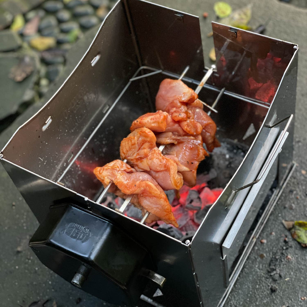 Marinated chicken skewers cooking inside a ROTO-Q 360 foldable grill over hot charcoal.